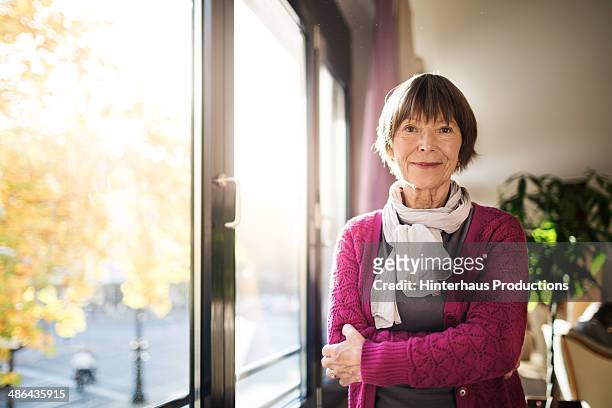 senior woman standing by window - mujer mayor fotografías e imágenes de stock