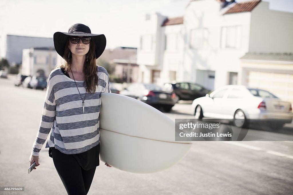 Girl in black hat walking w/surfboard & cell phone