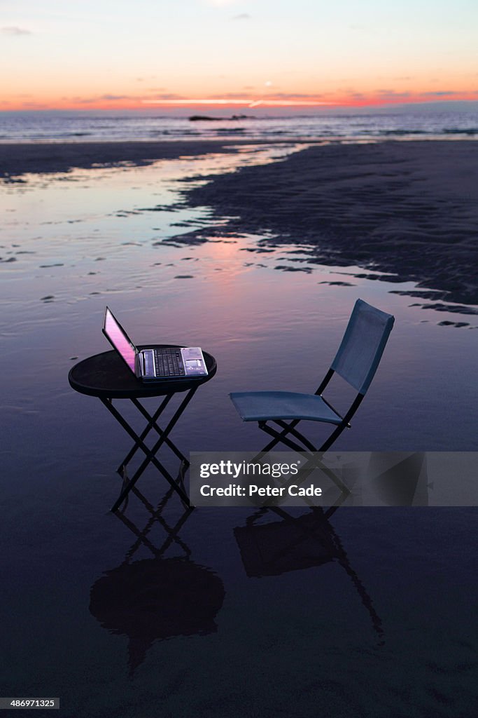 Table, chair and laptop on beach at sunset