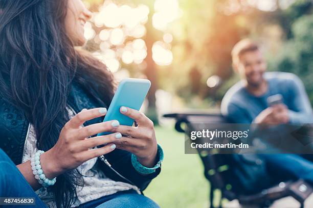 flirting couple in the park texting on smartphones - bokeh love stock pictures, royalty-free photos & images