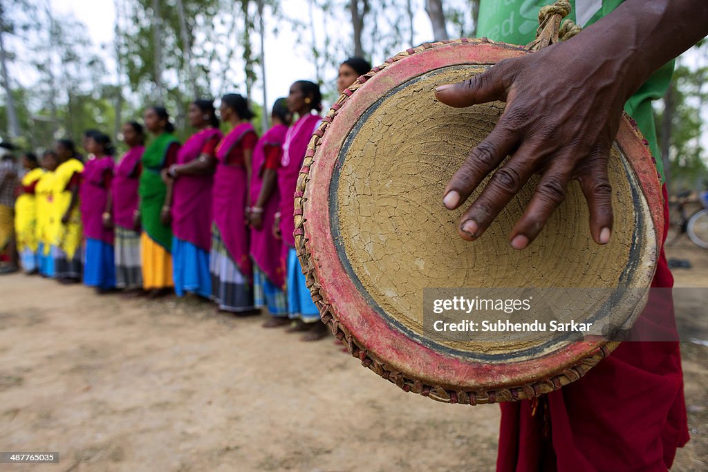 A Santhal man beats a drum as women dance in the background...
