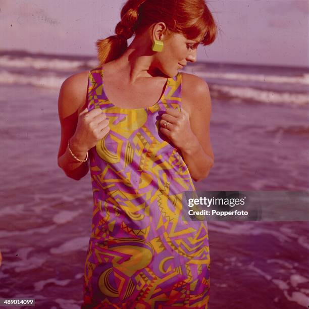 Sixties Fashion - A young female model is pictured wearing a patterned silky dress in a Pucci style print fabric as she poses on a beach next to the...