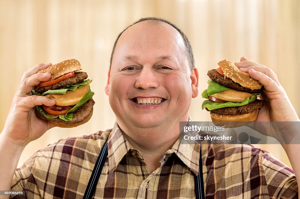 Happy fat man holding two hamburgers and looking at camera.