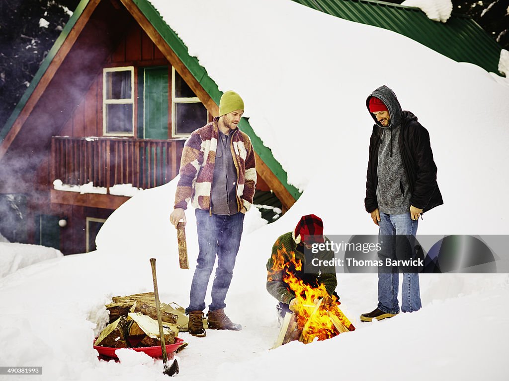 Three men building fire in snow in front of cabin