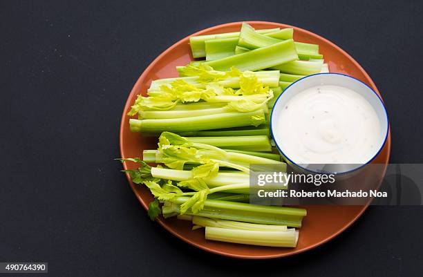 Celery sticks with leaves on few of them and a bowl full of delicious ranch sauce, placed in a brown plate against a black background.