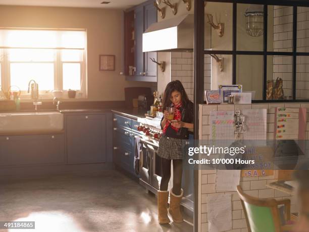 girl standing in a sunny kitchen looking at phone - sun flare on glass stockfoto's en -beelden