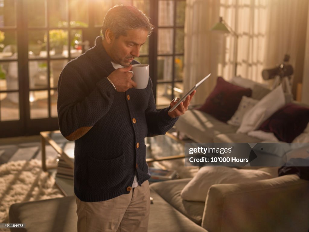 A man at home holding a cup and looking an tablet computer