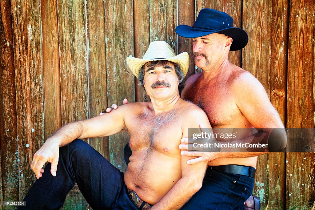 Two hairy bear affectionate cowboys against wooden fence