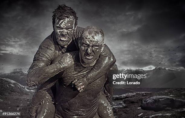 shaved man carrying friend during a mud run - manage stockfoto's en -beelden