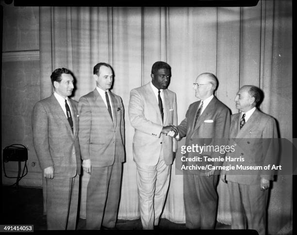 Group portrait of Al Levy, Louis A Radelat, Jackie Robinson shaking hands with John Maloney, and Bert Stern, standing in front of curtain,...