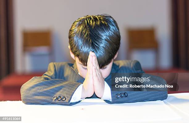 Young Christian boy praying in Catholic church during his first communion. He has straight dark hair, and kneels in front of a white altar with his...