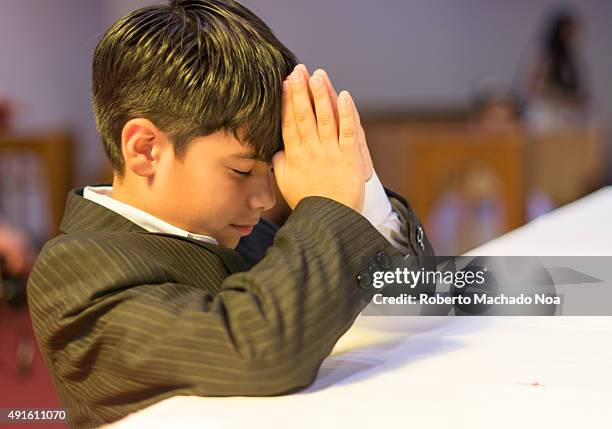 Young Christian boy praying in Catholic church during his first communion. He has straight dark hair, and kneels in front of a white altar with his...