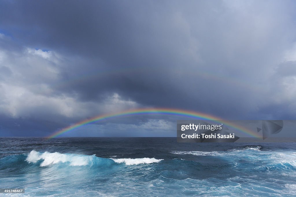 Rainbow appears over wave and horizon at dusk