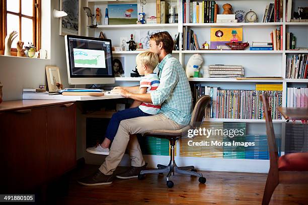 boy sitting on father's lap at computer desk - home office chair stock pictures, royalty-free photos & images