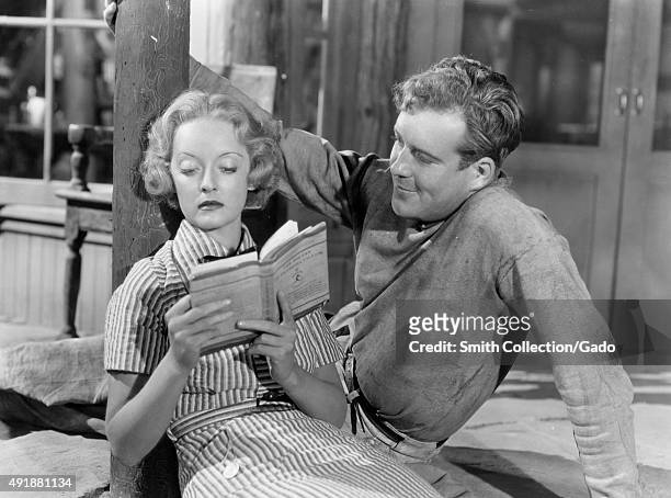 Bette Davis reclining on the floor and reading a book to a man, in a movie still, 1949.