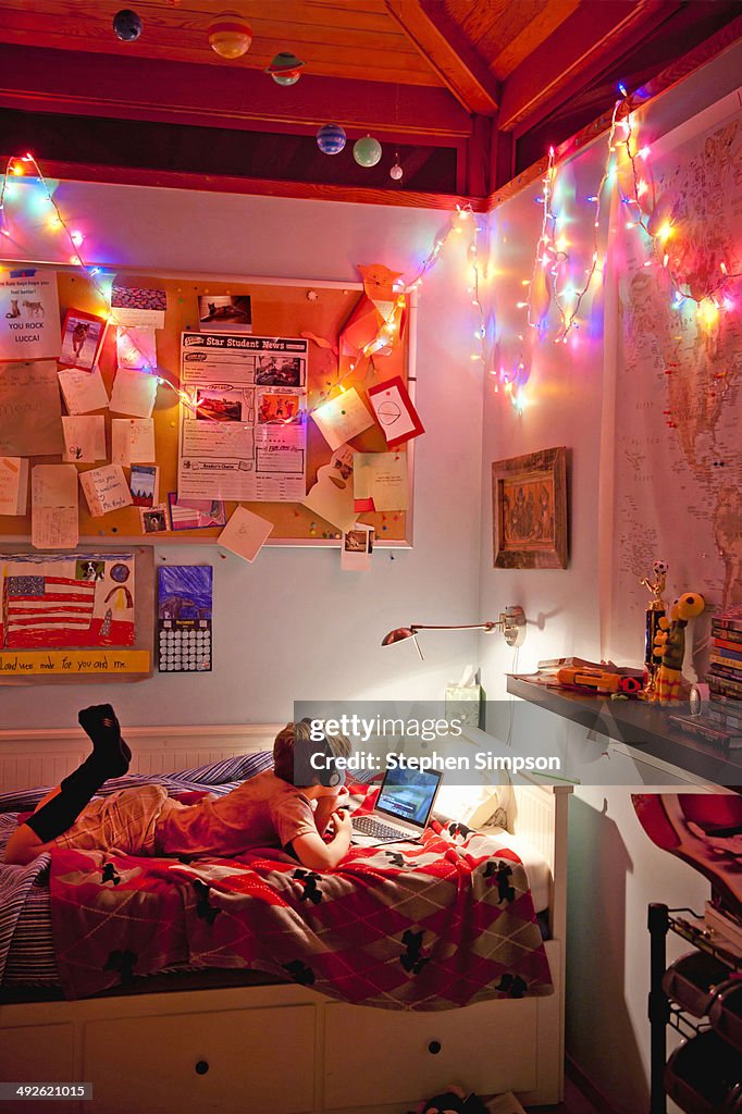 Boy with laptop on his bed, messy room with lights