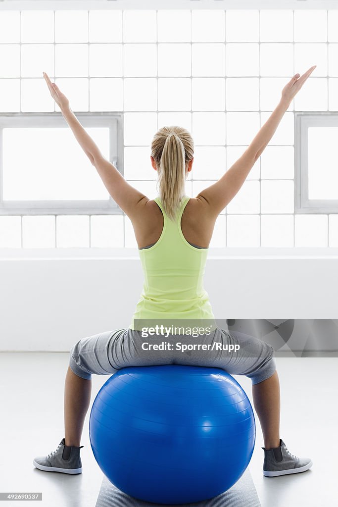 Young woman sitting on exercise ball, arms raised, rear view