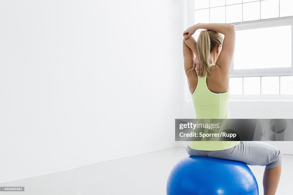 Young woman sitting on exercise ball, rear view, stretching