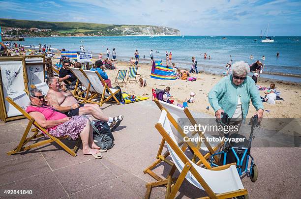 senior tourists enjoying sunshine deckchairs at busy seaside beach england - fat man tanning stock pictures, royalty-free photos & images