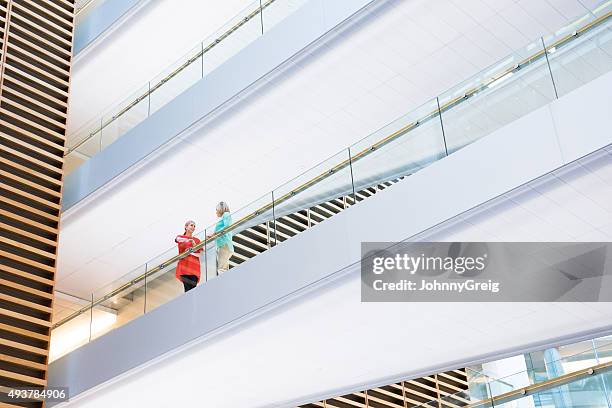 two businesswomen on overhead walkway in modern office building - johnny stark stock pictures, royalty-free photos & images