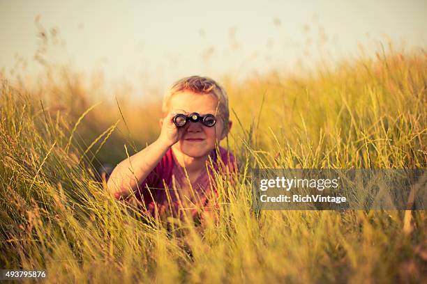 young boy looking through binoculars hiding in grass - day telescope stock pictures, royalty-free photos & images
