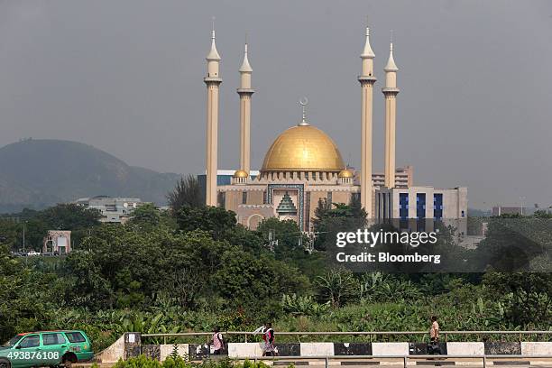 Pedestrians walk by as the minarets of the Central Mosque stand in the distance in Abuja, Nigeria, on Wednesday, Oct. 21, 2015. A drop in crude...