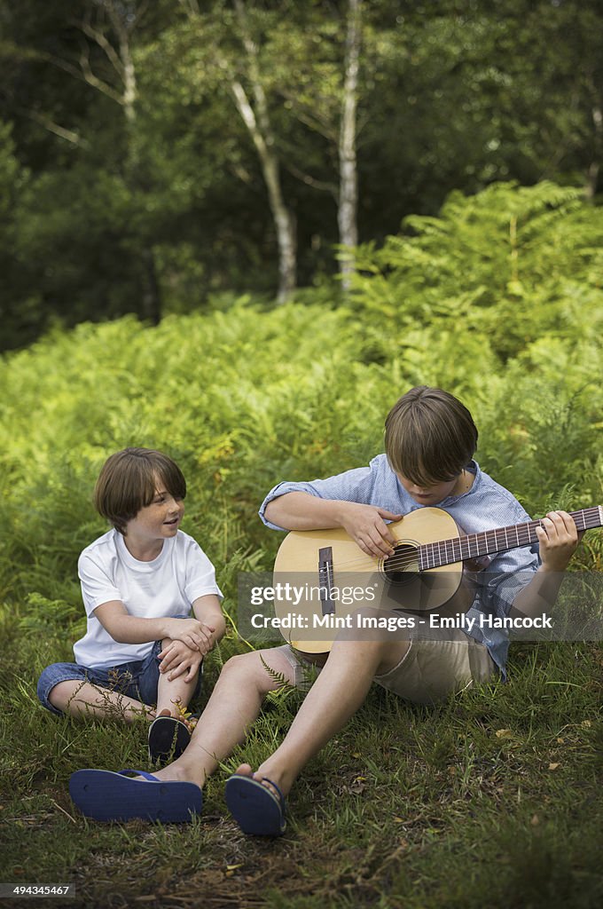 Two boys camping in New Forest. Sitting on the grass, one playing a guitar.