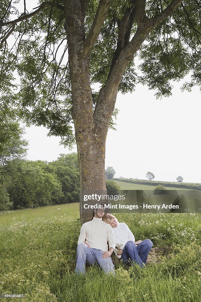 A couple sitting leaning against a tree in a meadow.