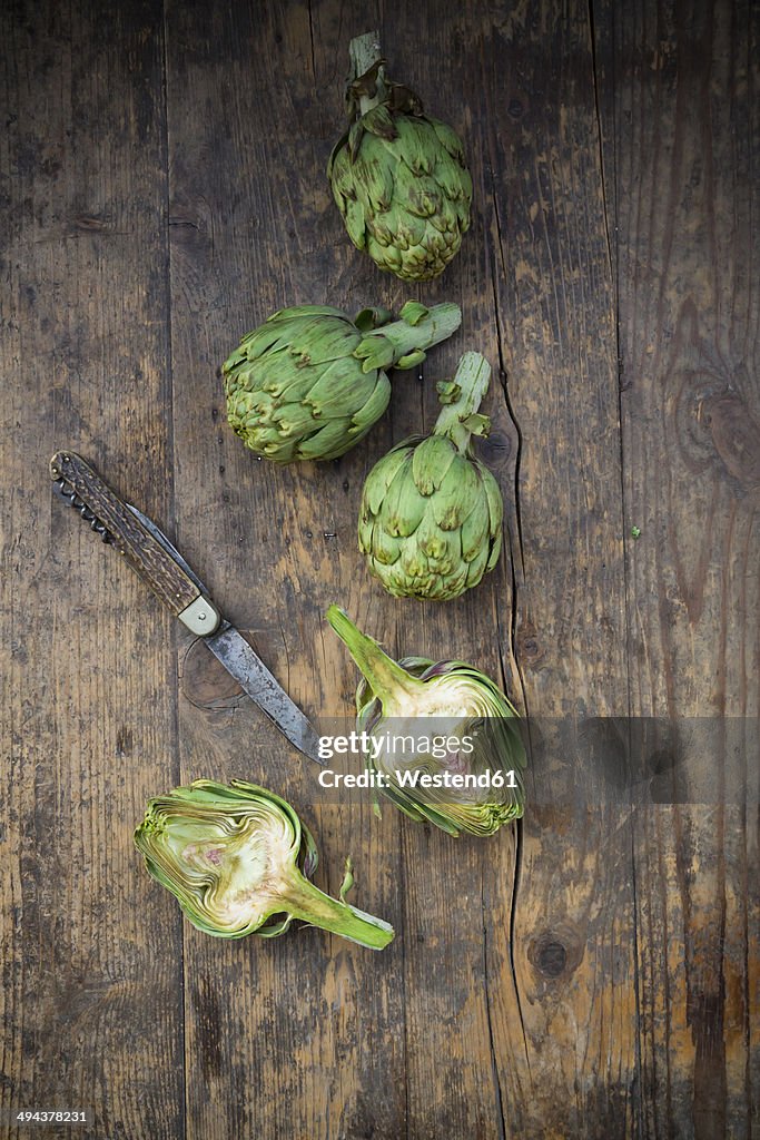 Sliced and whole organic artichokes and kitchen knife on wooden table, view from above