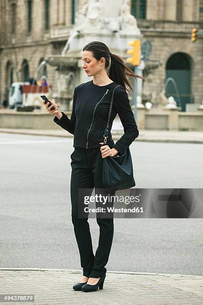 spain, catalunya, barcelona, young black dressed businesswoman looking at her smartphone in front of a street - calling on the side road stock pictures, royalty-free photos & images