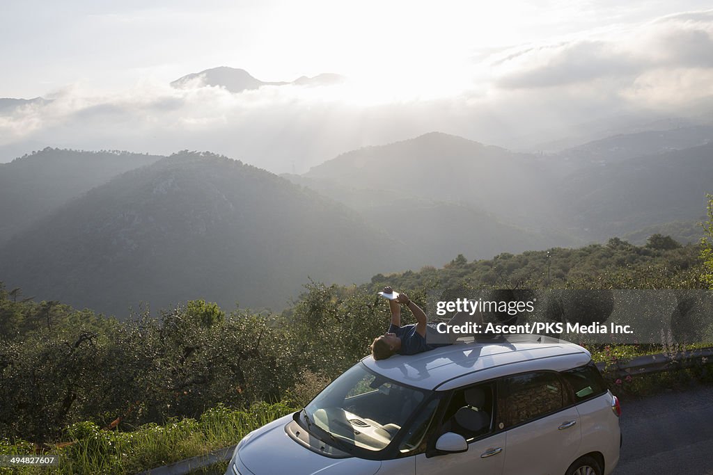 Young man lies on roof of car, uses digital tablet