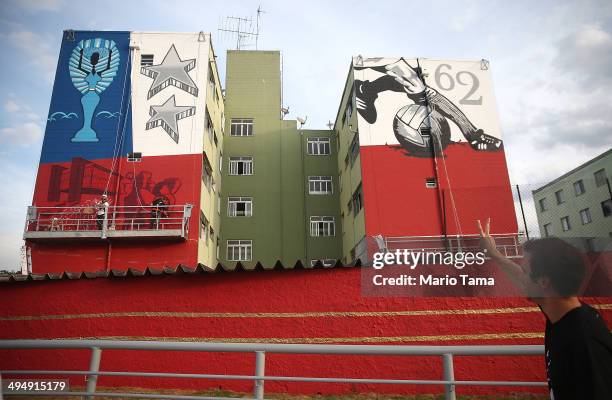 Man flashes the 'peace' sign walking past artists painting graffiti on the side of a building honoring Brazil's 1962 World Cup victory over...
