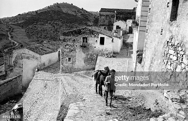 Suggestive view of the village of Rocca Imperiale with the hills on the background; a boy with a donkey is climbing the steep road going to the...