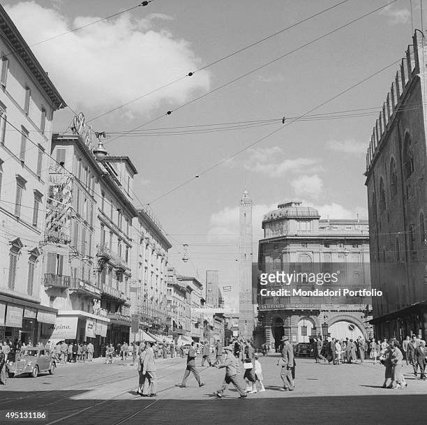 Some people walking on Piazza Maggiore and via Ricasoli. In the distance, the Torre Garisenda and the Torre degli Asinelli standing. Bologna, May 1955