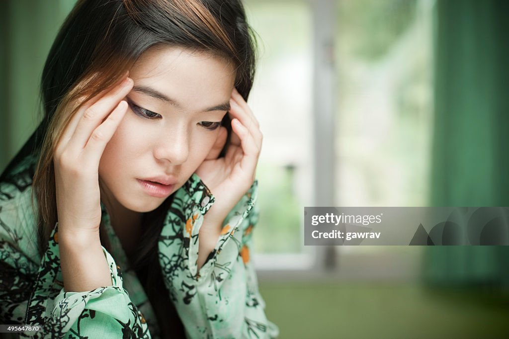 Sad Asian cute girl sitting near window holding her head.