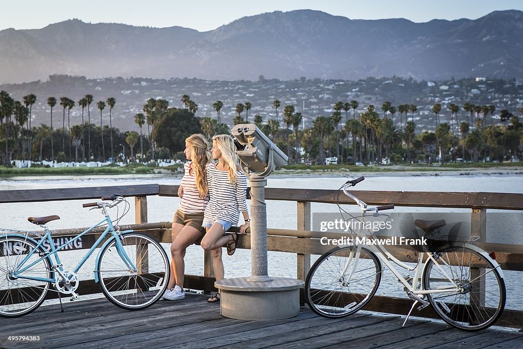 Two women in their twenties taking a break from riding their bikes. They are resting on the railing watching the sun go down with mountains and ocean in the background.