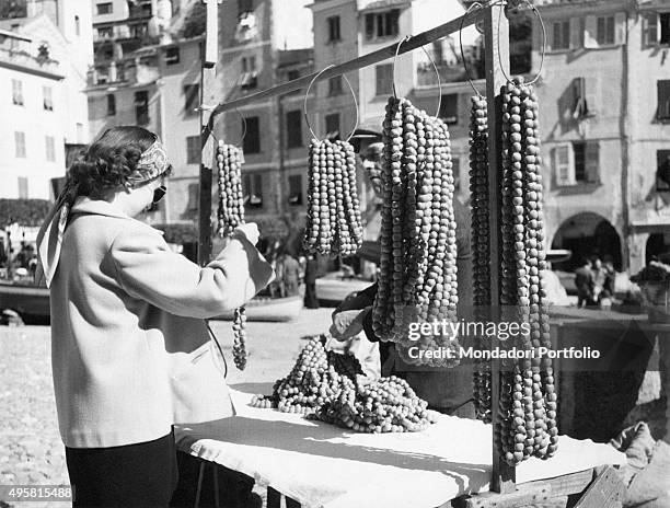 Tourist wearing sunglasses takes a look on hazelnuts necklaces of various sizes exposed in an outdoor stand. Portofino , Italy, 1960.