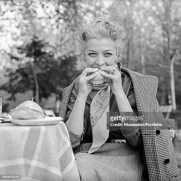 The actress Isa Barzizza bites a salami sandwich with taste, sitting at a small table in a park, with her overcoat placed on her shoulders. Italy,...