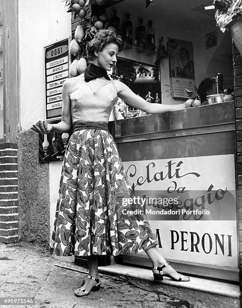 Model posing in front of an ice cream kiosk in a dress designed by the fashion house Antonelli. Italy, 1955