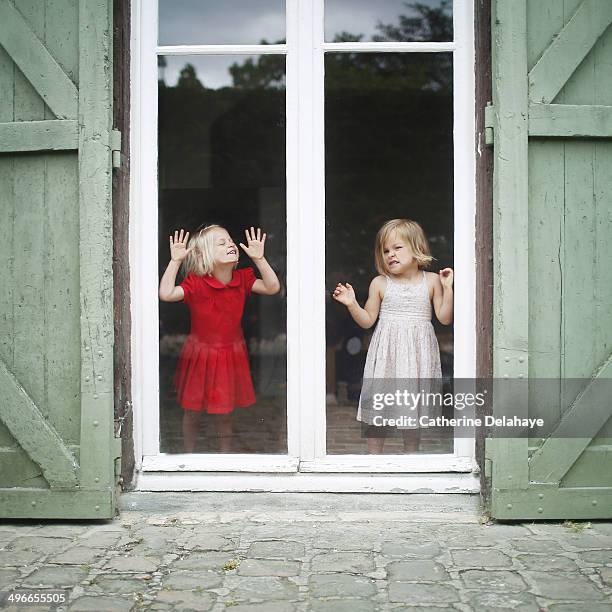 two 3 years old twins girls posing behind a window - een gek gezicht trekken stockfoto's en -beelden