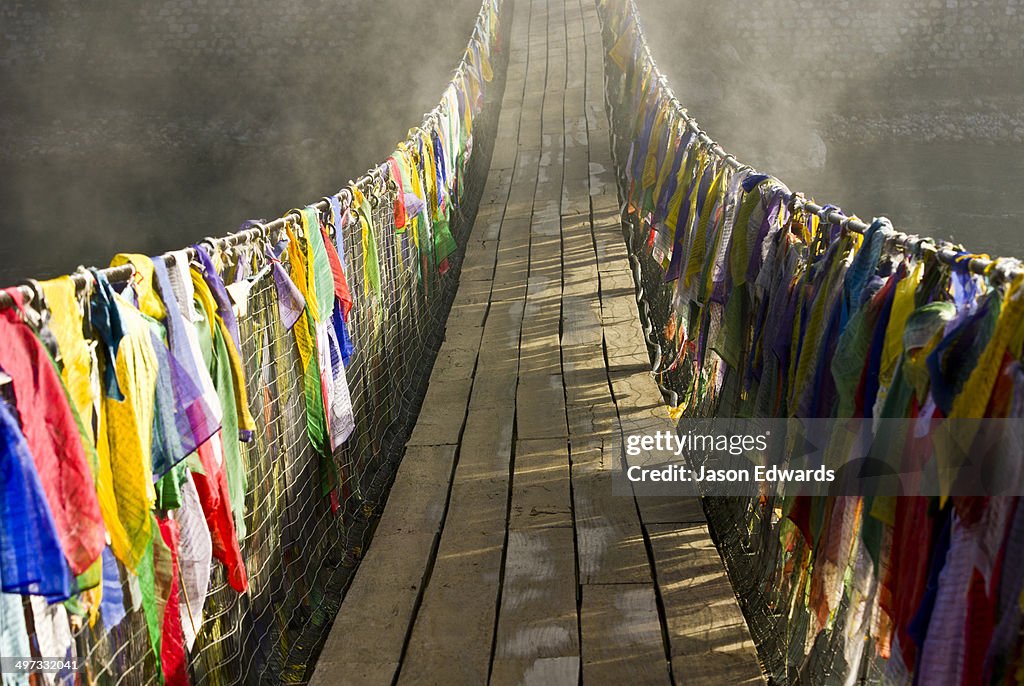 Prayer flags drape a suspension bridge across a river as dawn melts the fog away.