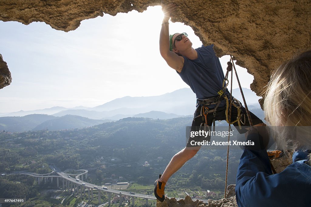 Climber stretches for hold on cave edge, hwy below