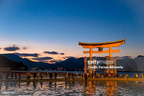 People walk past Itsukushima Shrine and its floating torii gate at sunset on a cold autumn evening.