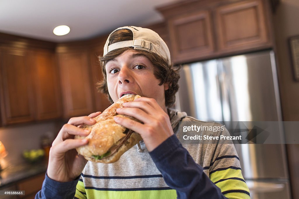 Teenage boy in kitchen biting large sandwich