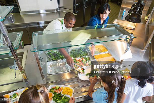 cafeteria worker serving trays of healthy food to children - kantine stockfoto's en -beelden