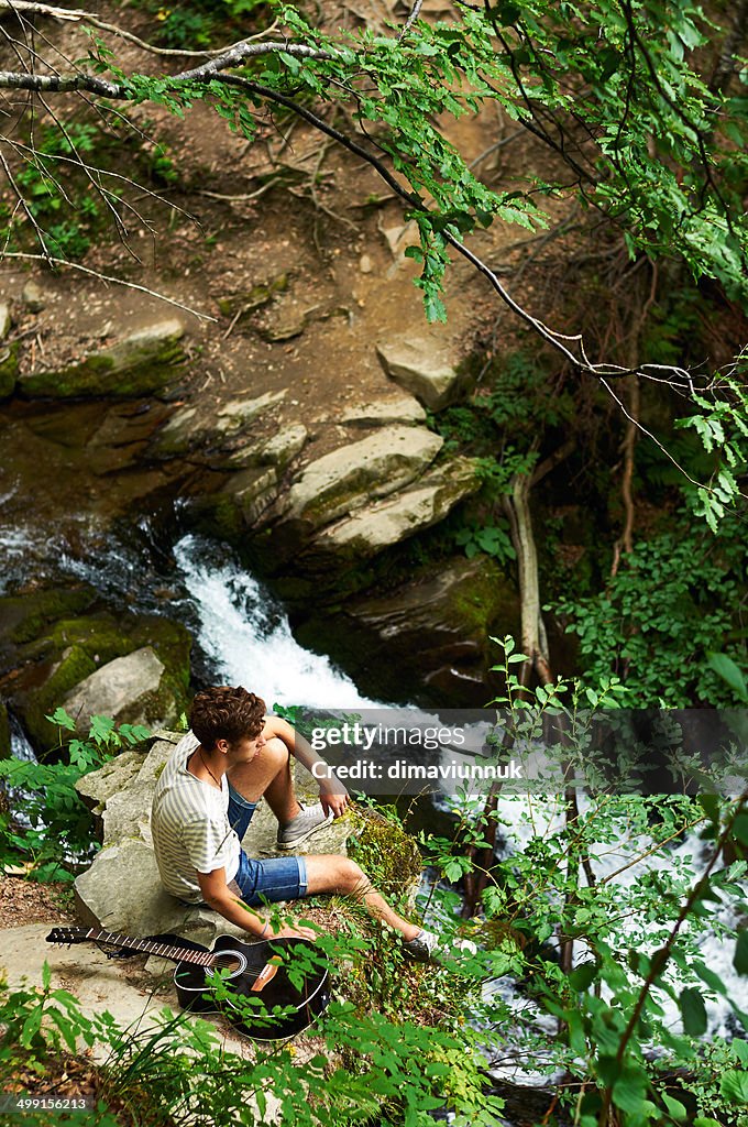 Elevated view of teenage boy (14-15) with guitar sitting on rock by river