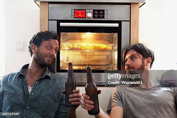 two men clinking beer bottles at oven in kitchen - male friends drinking beer stock pictures, royalty-free photos & images