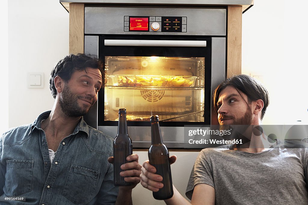 Two men clinking beer bottles at oven in kitchen