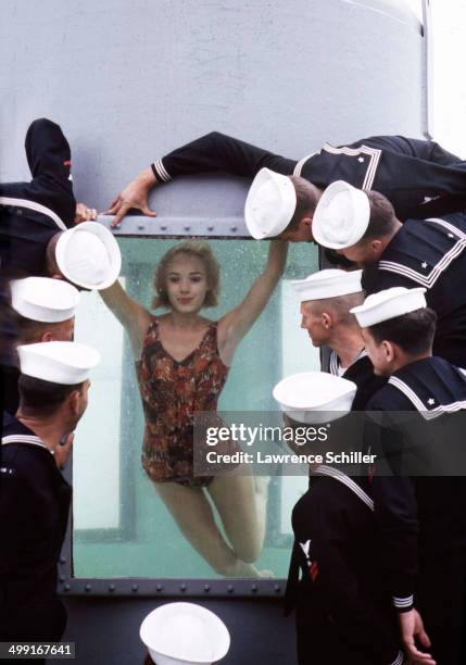 Group of navy sailors look at a model in a swimsuit as she poses, underwater, behind a window, 1963. The photograph was taken as part of a shoot for...