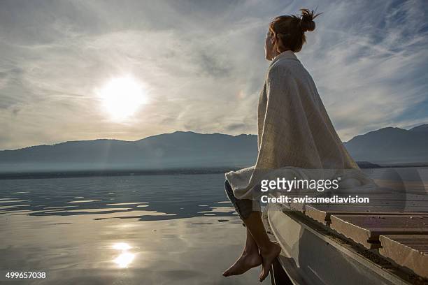 young woman relaxes on lake pier with blanket, watches sunset - tranquil scene stock pictures, royalty-free photos & images